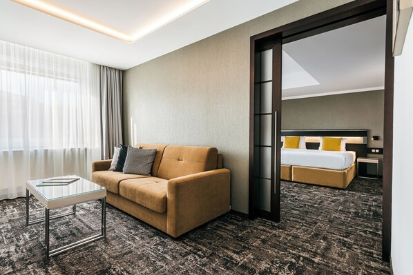 Habitación de hotel con sofá, una mesita de centro de vidrio, cama con almohadas amarillas y vistas a través de una puerta abierta.