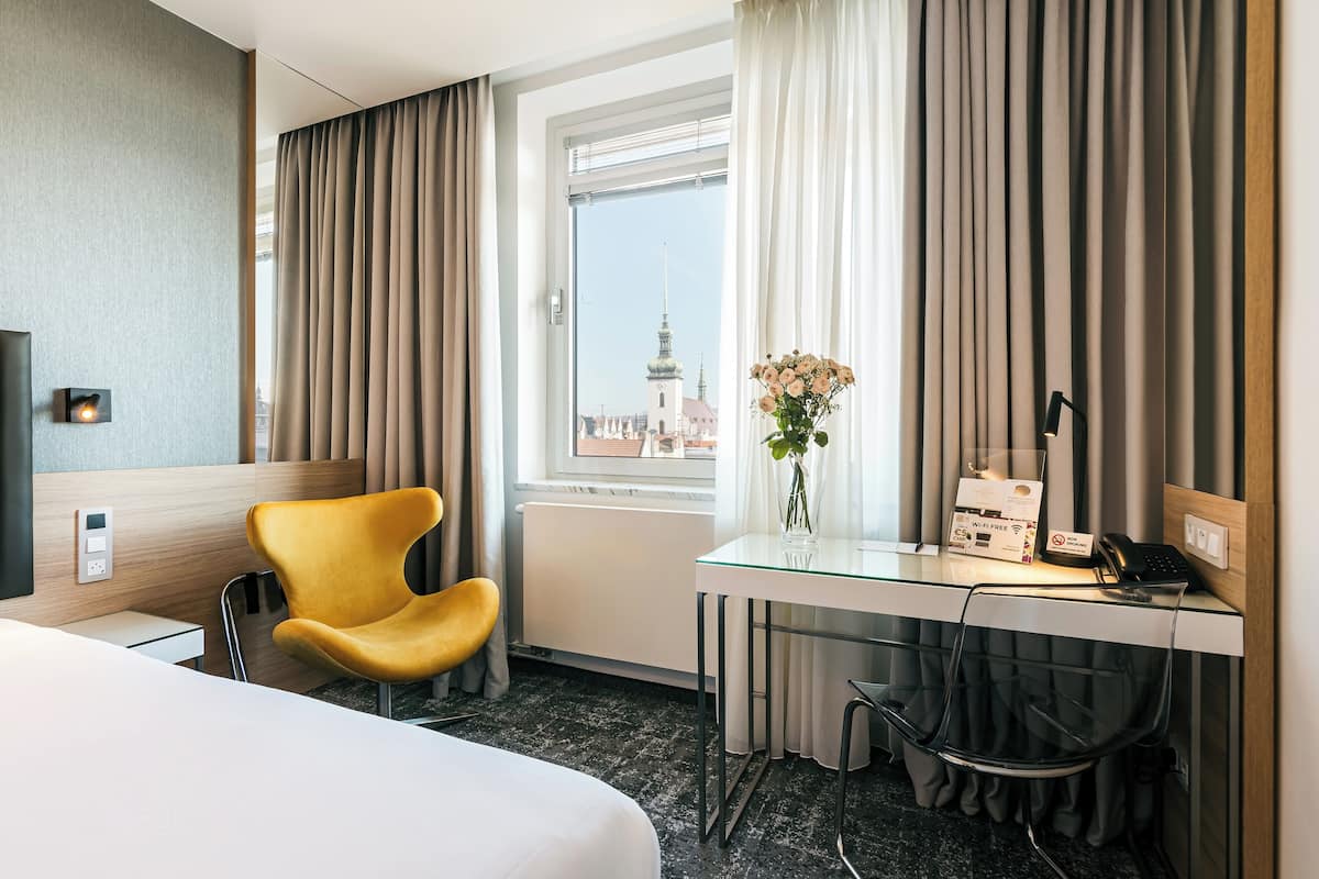 Hotellihuone, jossa on sänky, työpöytä tietokoneella, tuoli, maljakko kukkineen ja ikkunasta avautuva näkymä kaupunkimaisemaan.