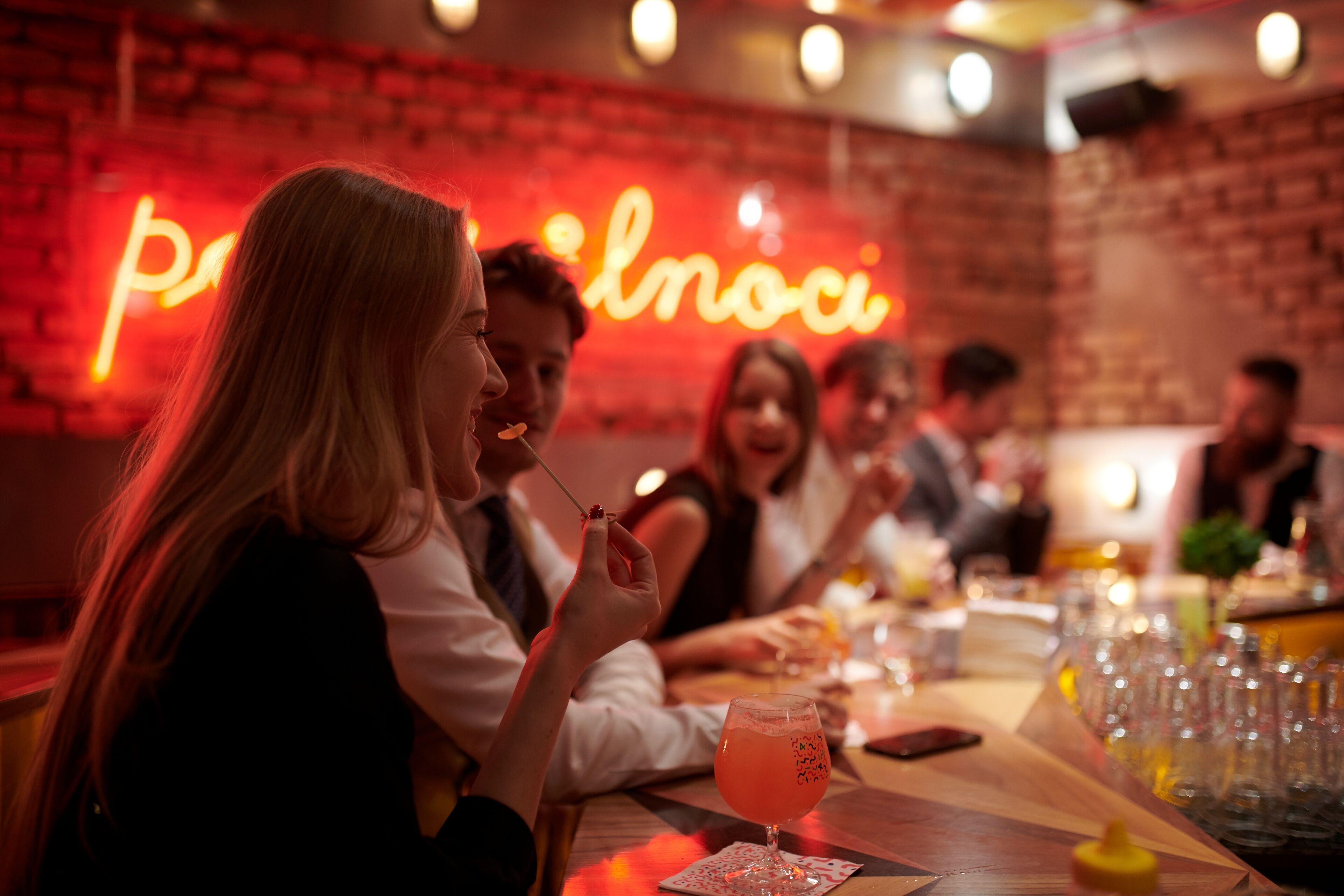 Eine Gruppe von Personen speist in einem Restaurant, im Hintergrund eine Ziegelwand und ein Neonzeichen.