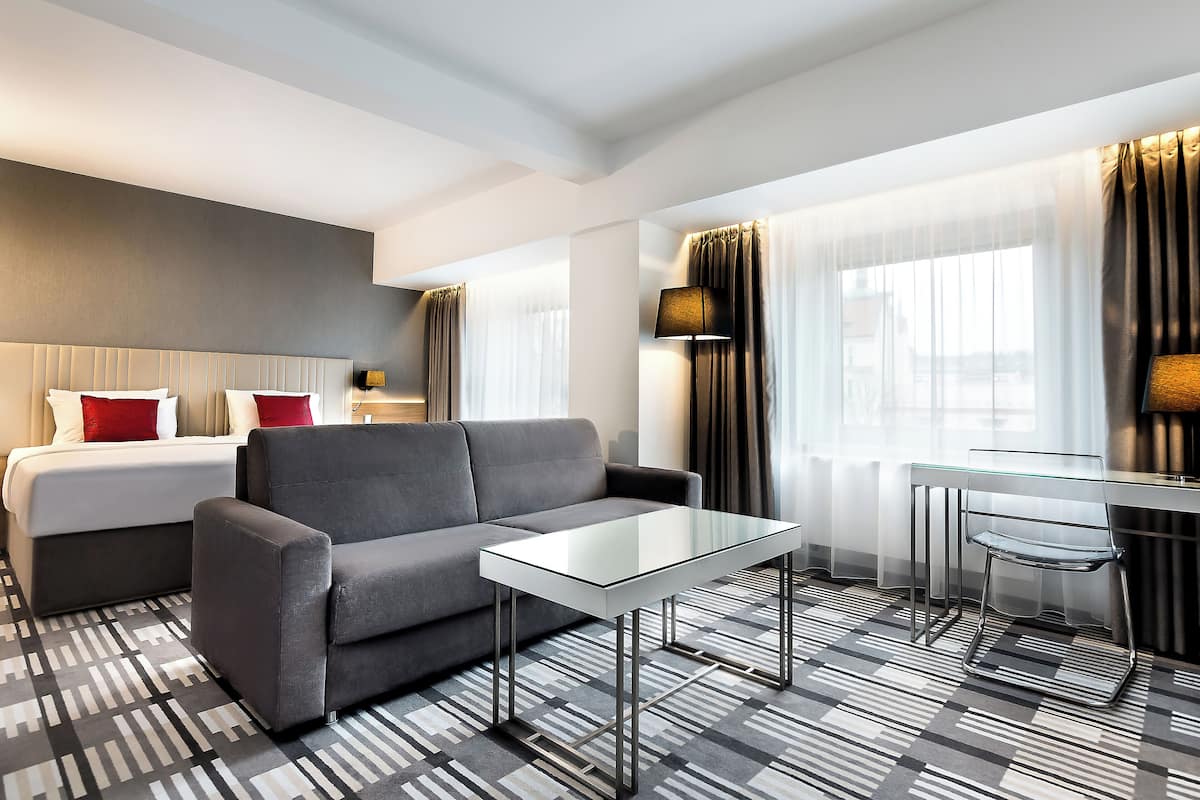 Moderni hotellihuone, jossa on sänky, harmaa sohva, lasipöytä ja pieni ruokailutila.