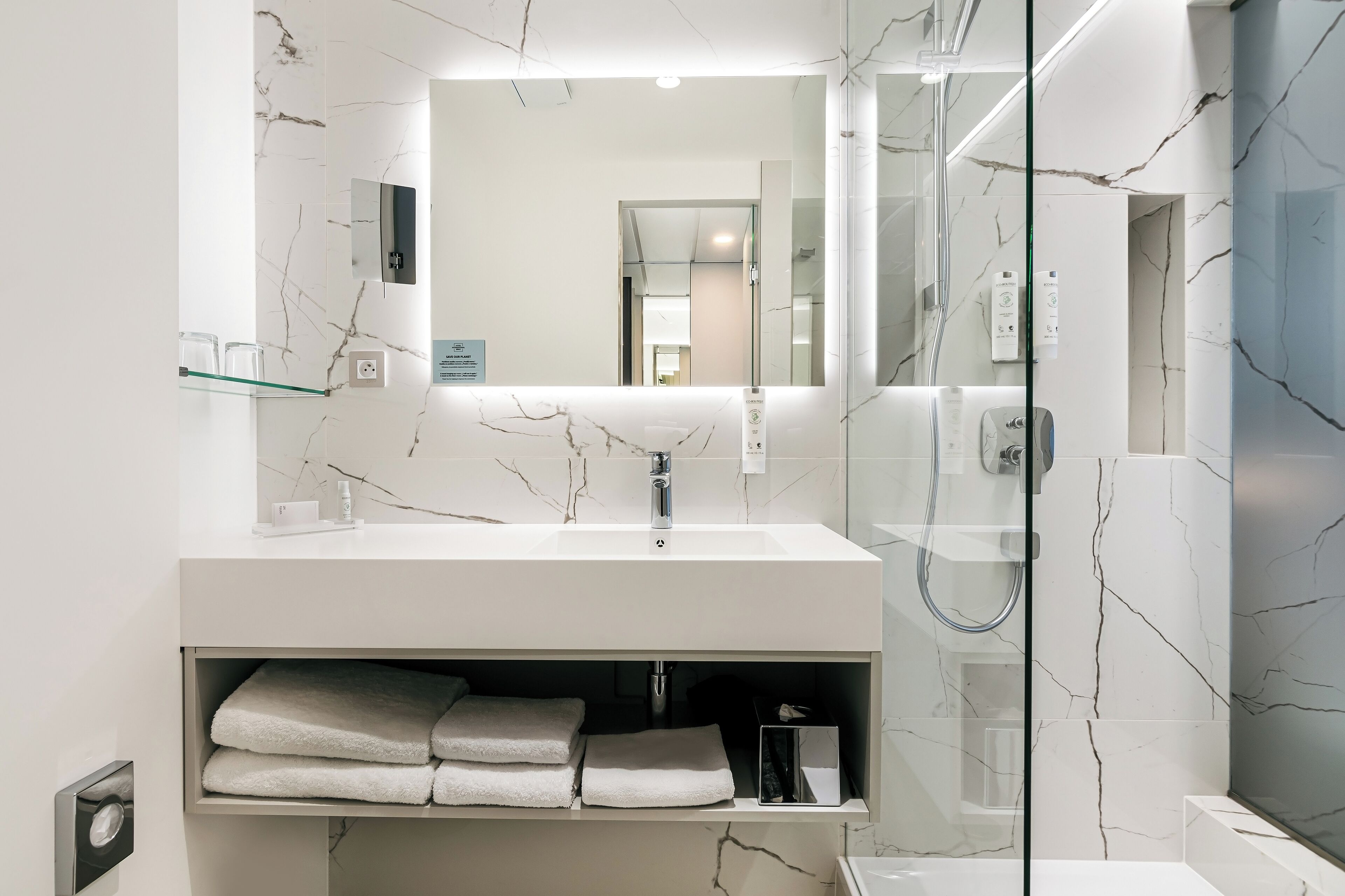 Un baño moderno con salpicadero de mármol, un espejo grande, un lavabo blanco y una ducha con mampara de vidrio.