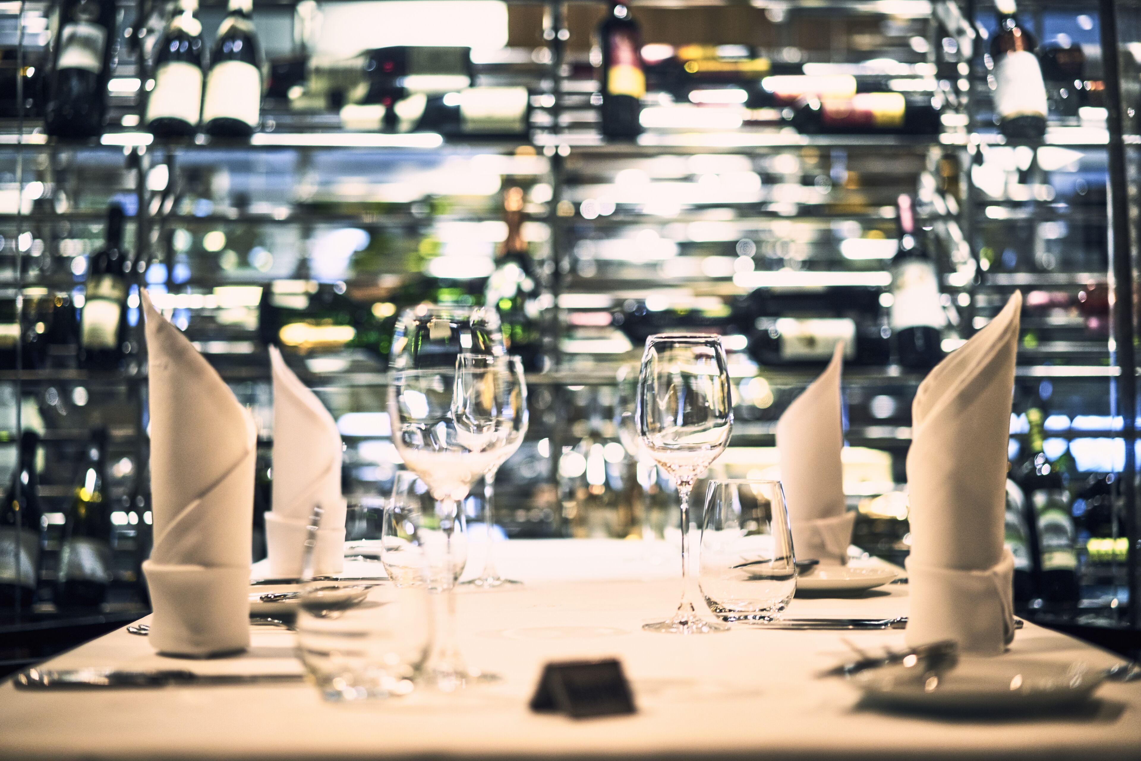 A table set with wine glasses and napkins, with a backdrop of wine bottles.