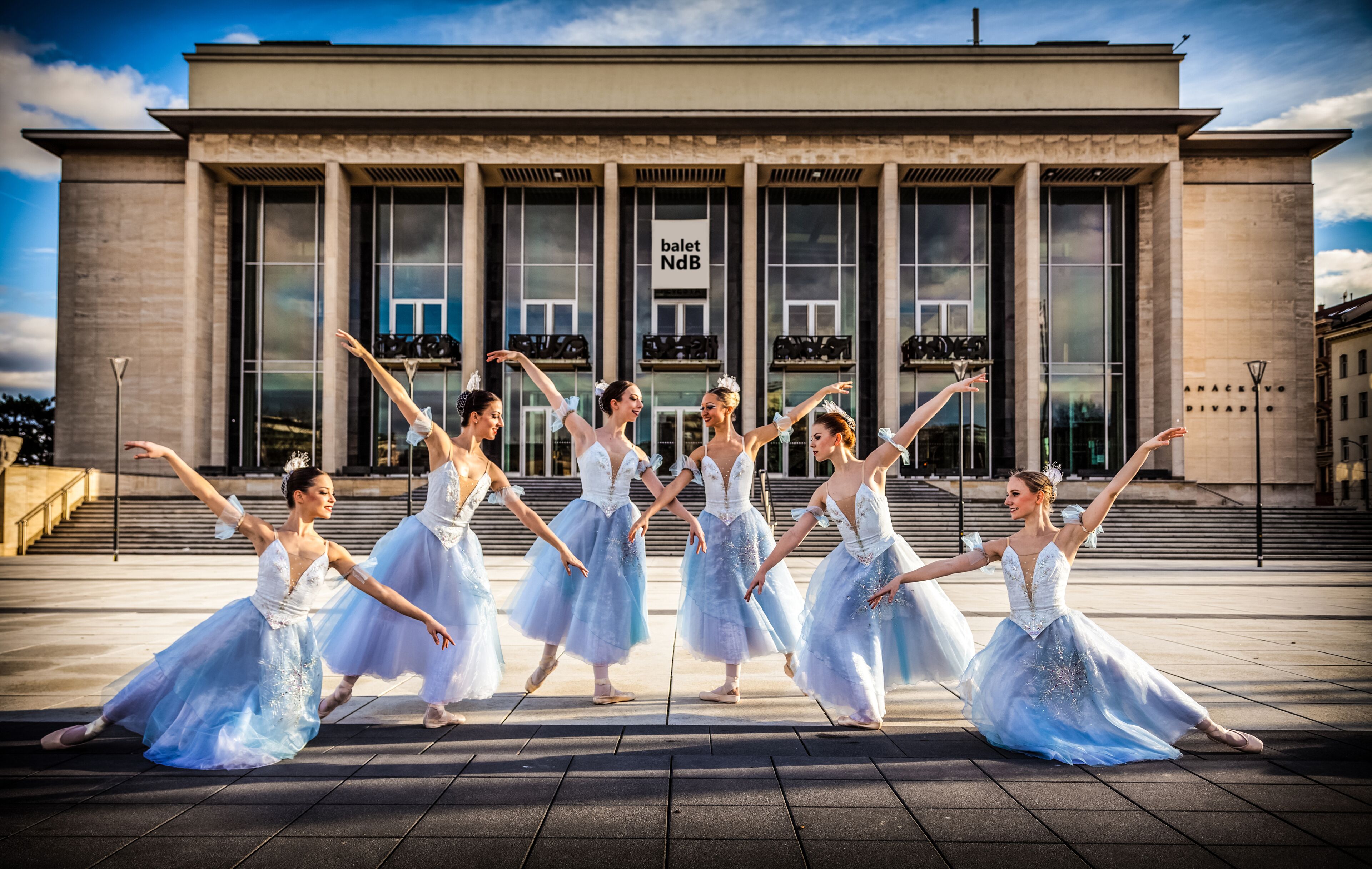 Un grupo de bailarines de ballet actuando frente a un edificio con grandes ventanales y un cartel que dice 'ballet NdB'.
