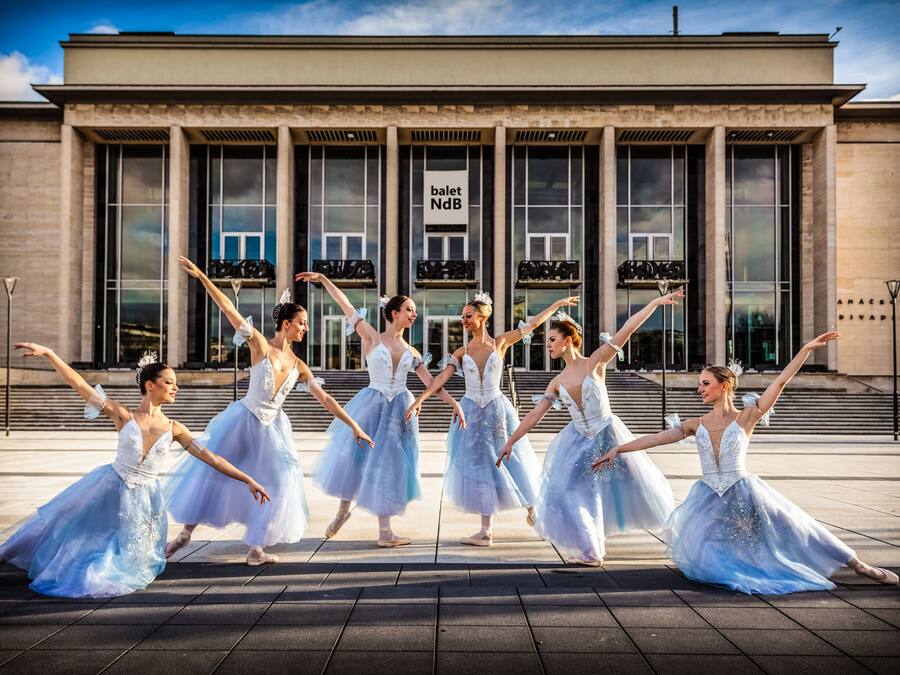 Un grupo de bailarines de ballet actuando frente a un edificio con grandes ventanales y un cartel que dice 'ballet NdB'.