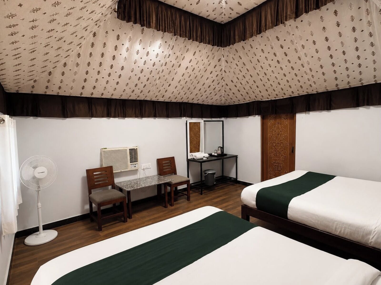 Ein Hotelzimmer mit zwei Betten, einem Schreibtisch mit Stuhl, einem Ventilator und einem Spiegel.