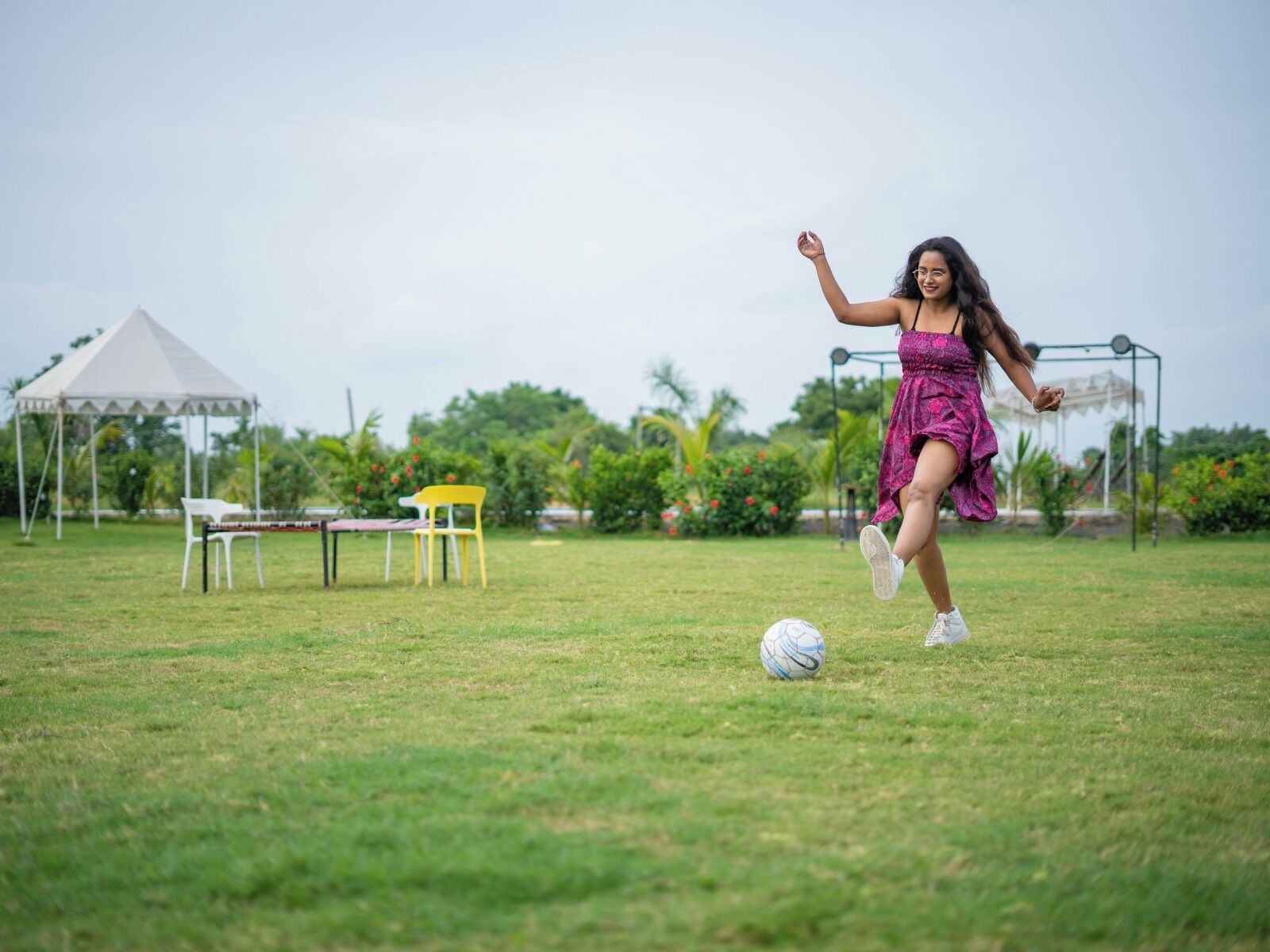Uma pessoa jogando futebol em um campo gramado, com uma barraca e cadeiras ao fundo.