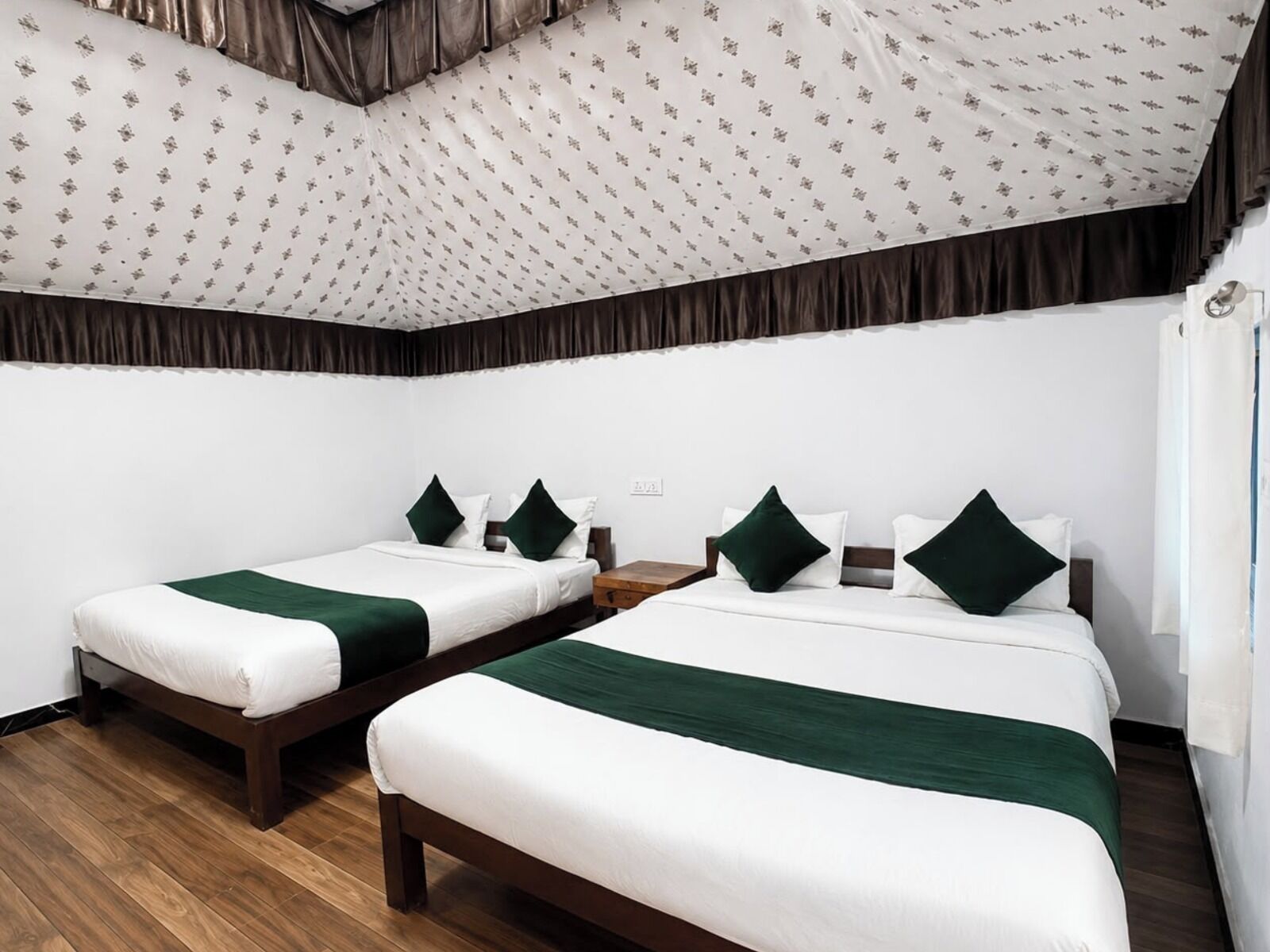 A hotel room with two single beds, each with a green accent pillow, a wooden nightstand, and a patterned ceiling.