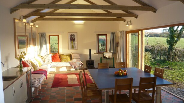 A cozy room with a bed, a dining table with chairs, a kitchen area, and large windows offering a view of the outdoors. - Swallow cottage (Kilgetty)