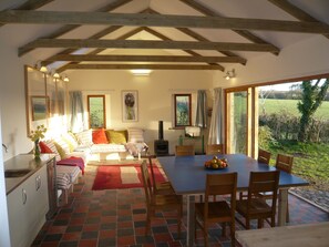 A cozy room with a bed, a dining table with chairs, a kitchen area, and large windows offering a view of the outdoors. - Swallow cottage (Kilgetty)