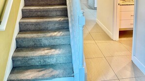 Una scala con gradini rivestiti di moquette e ringhiere bianche, che conduce a una stanza con una porta bianca e una zona cucina visibile sullo sfondo.