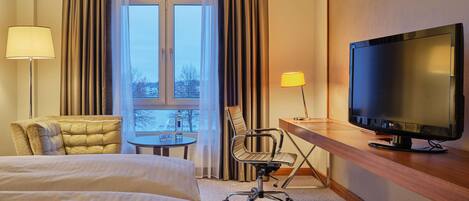 A hotel room with a bed, a desk with a television, a chair, a table with glasses, and curtains.