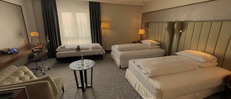A hotel room with two single beds, a double bed, a desk with a laptop, a chair, a small table with glasses, and a lamp.