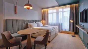Una habitación de hotel moderna con cama, una mesa redonda con sillas, televisión y un ventanal con cortinas.