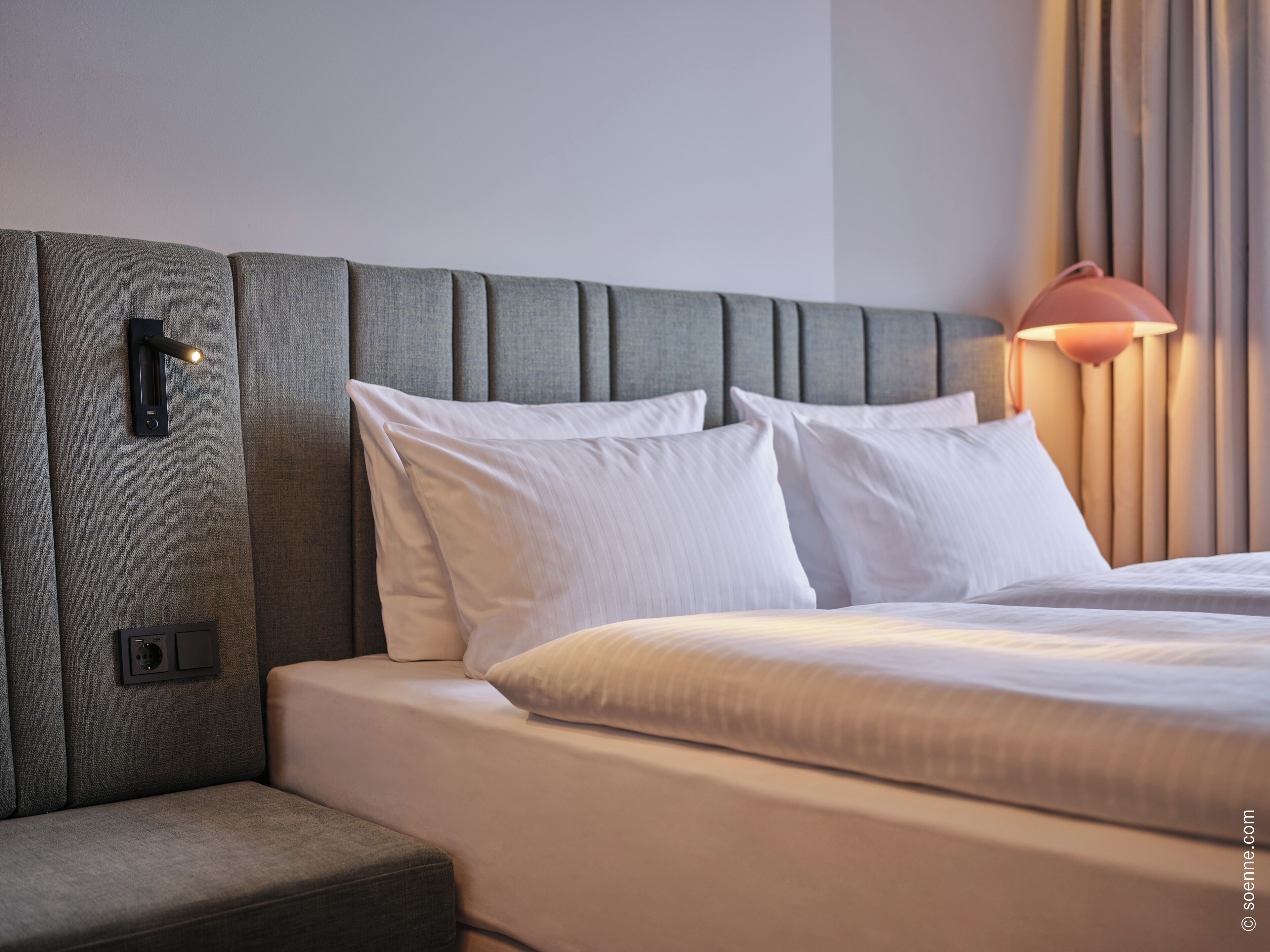 A neatly made bed with white linens, a grey upholstered headboard, and a bedside lamp.