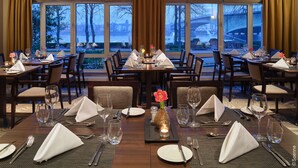 A restaurant with multiple tables set for dining, large windows with a view of a cityscape, and a warm interior lighting. - Dorint Hotel Bonn (Bonn)