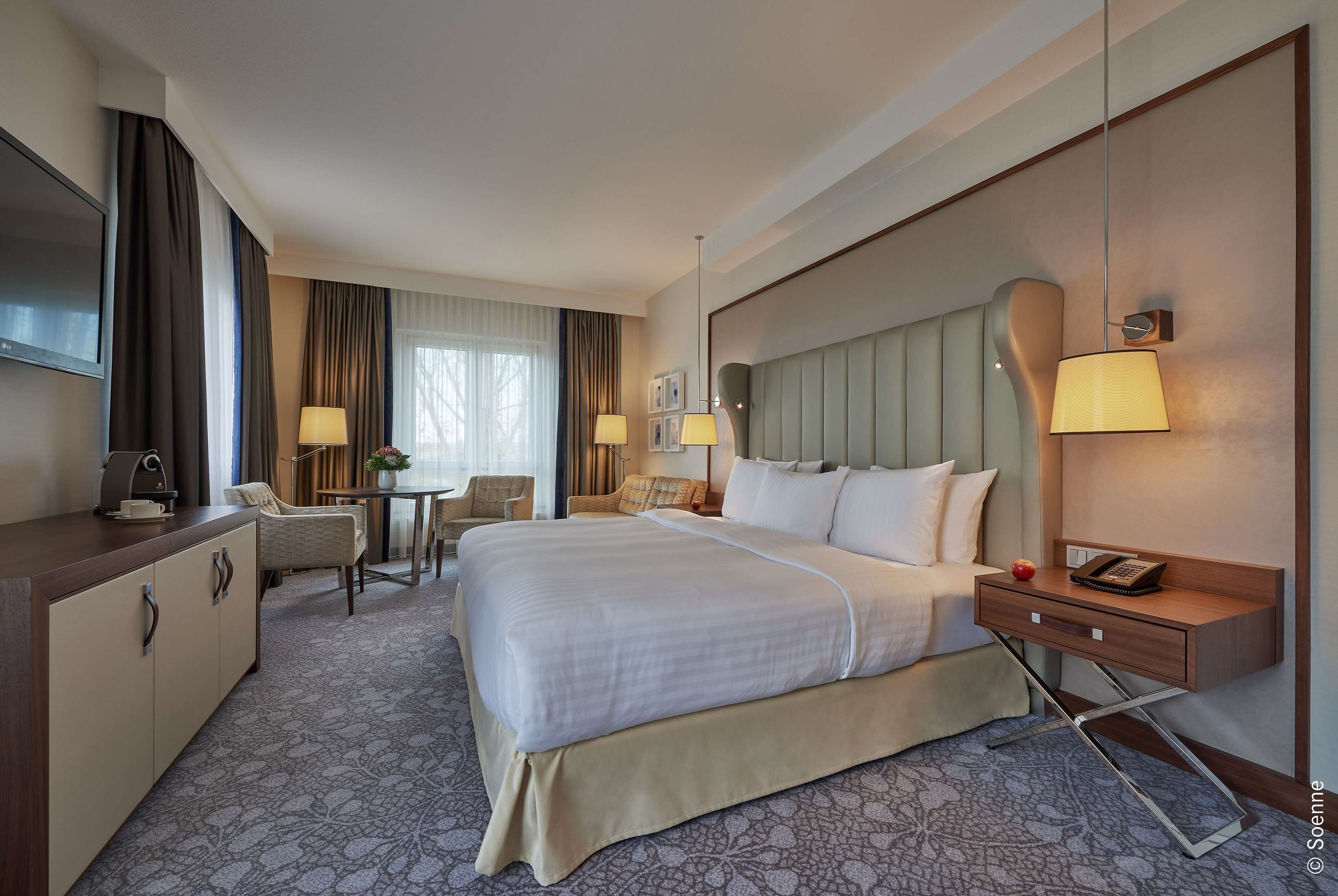 A hotel room with a large bed, a desk with a lamp, a chair, a TV, and a bedside table with a phone.