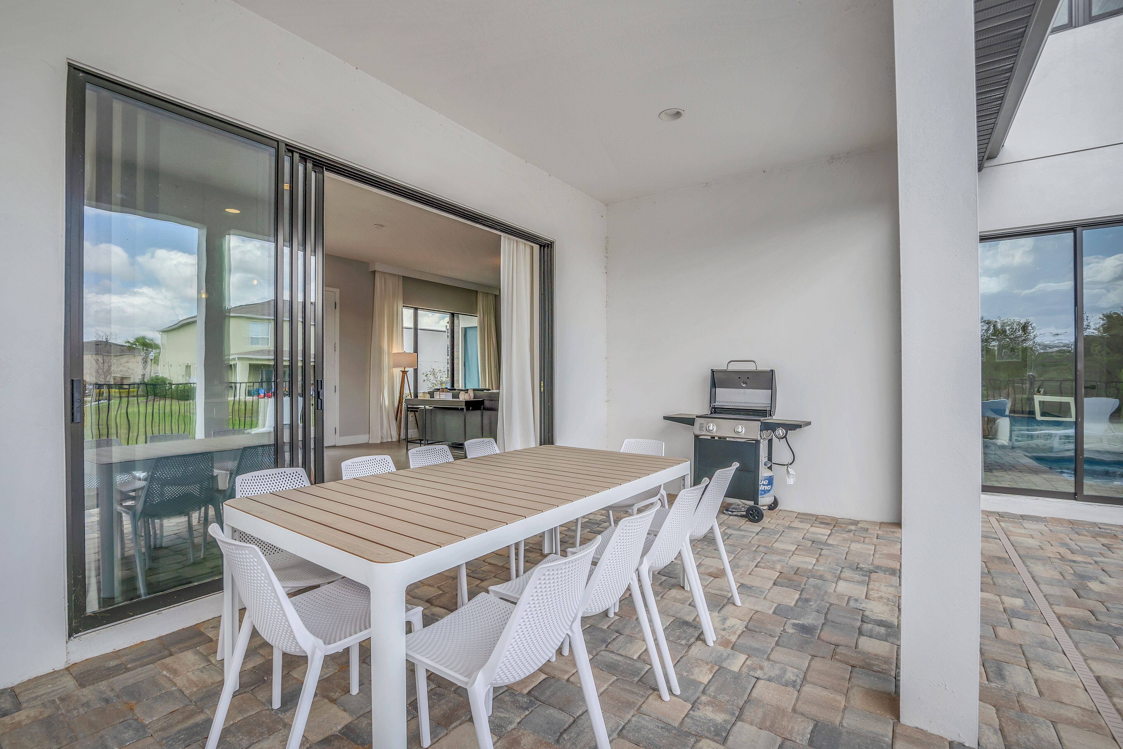 An outdoor patio with a wooden table and white chairs, a grill, and a view of a swimming pool.