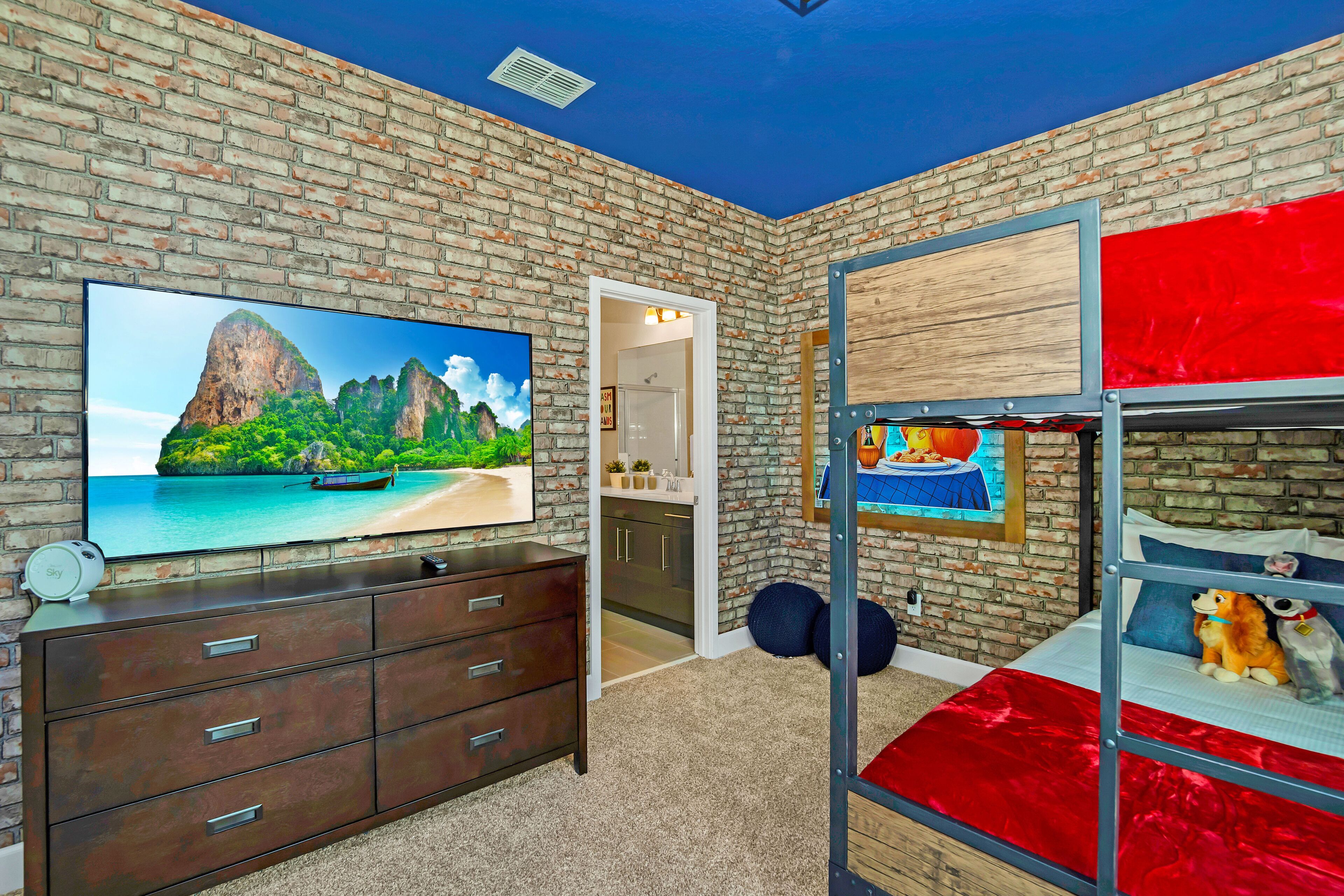 Une pièce avec un mur en briques, un téléviseur posé sur une commode, un lit superposé avec un matelas rouge et une porte donnant sur une salle de bain.