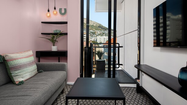 A modern living room with a sofa, a coffee table, and a TV mounted on the wall. There are shelves with decorative items and a potted plant. A sliding door leads to a balcony with a view of buildings and a mountain. - Miami Living at The Flamingo! (Cape Town)