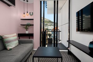 Una sala de estar moderna con un sofá, una mesa de centro y un televisor montado en la pared. Hay estantes con objetos decorativos y una planta en maceta. Una puerta corredera da a un balcón con vistas a edificios y una montaña.