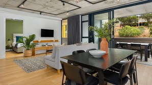 A modern living room with a sofa, dining table, and chairs. There is a TV mounted on the wall, a plant, and a window with curtains. - Modern 3-Bedroom in the Heart of Bo-Kaap - Netflix (Cape Town)