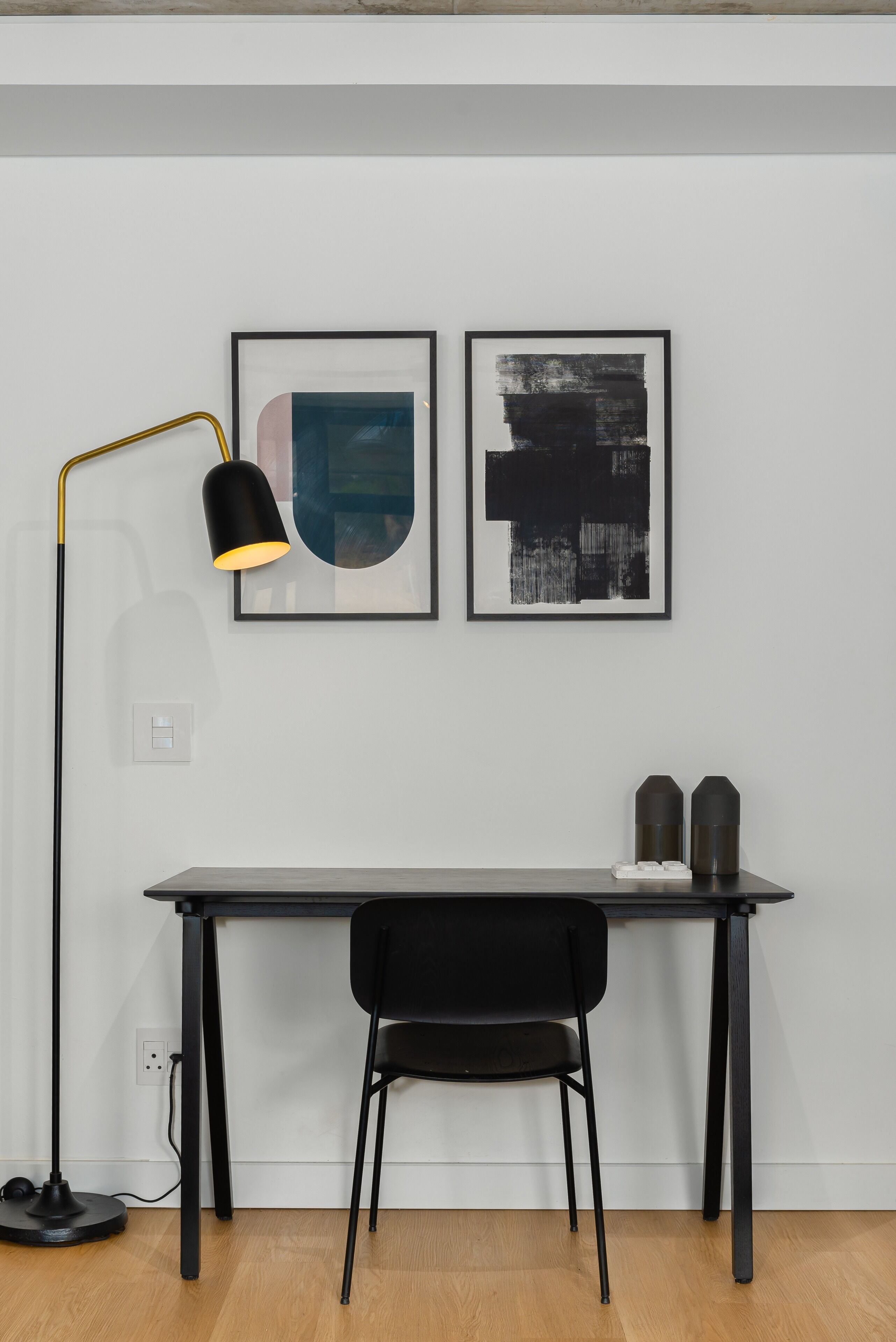 Un espace de bureau moderne comprenant un bureau, deux chaises, une lampe sur pied et deux tableaux abstraits encadrés accrochés au mur.