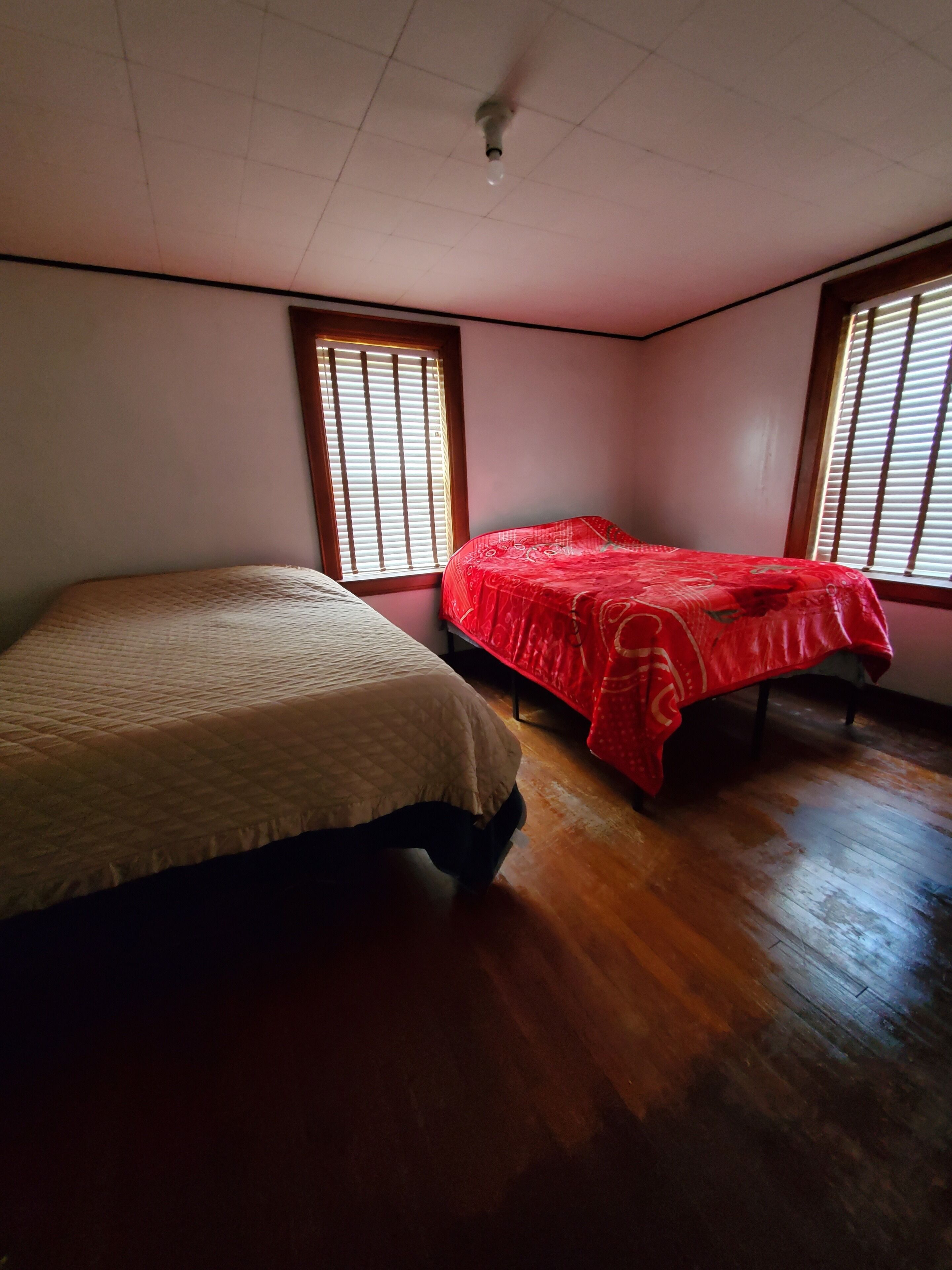 A room with two beds, wooden flooring, and windows with blinds.