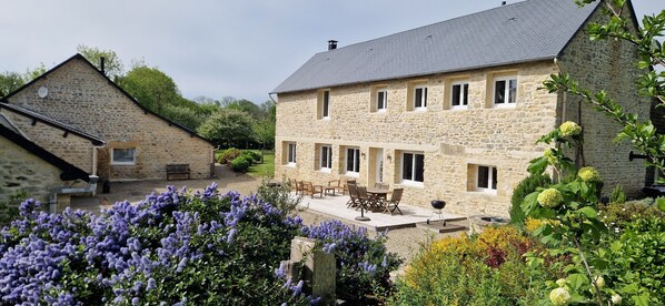 Un bâtiment en pierre doté d’un patio, d’un espace extérieur aménagé pour s’asseoir et d’un jardin où poussent des fleurs violettes.