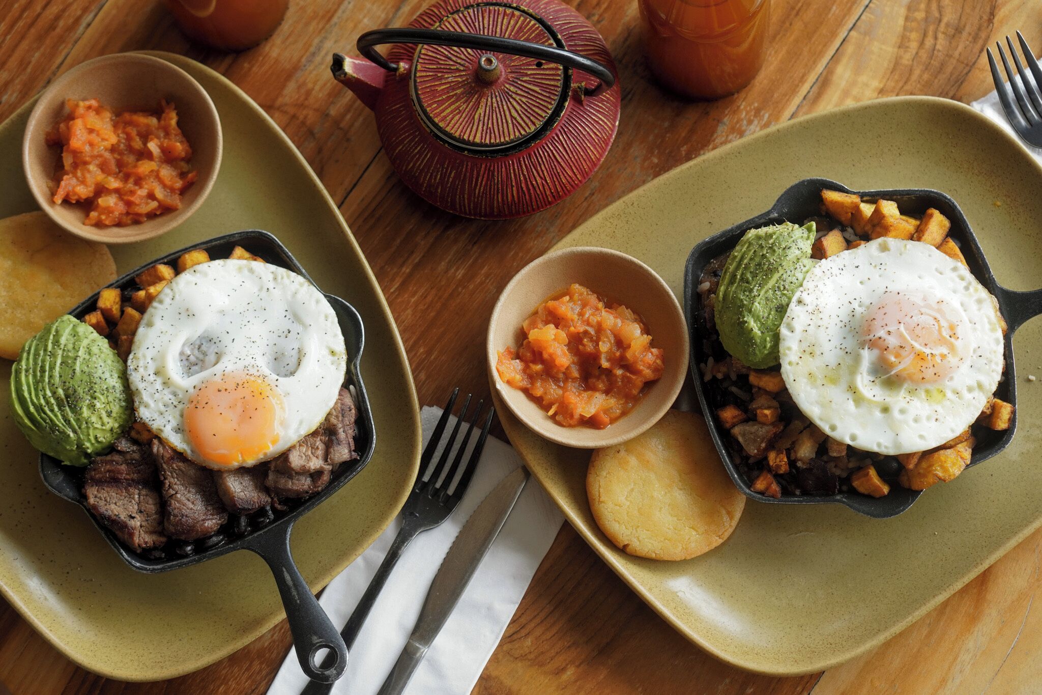 Une table dressée pour le petit-déjeuner, avec deux poêles contenant des œufs et de la viande, accompagnées de bols de salsa et d’avocat.