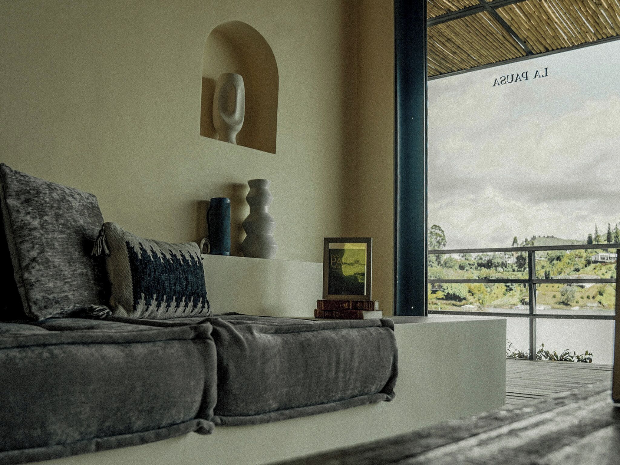Una sala de estar moderna con un sofá gris, una mesita con adornos y un amplio ventanal con vistas a la vegetación.