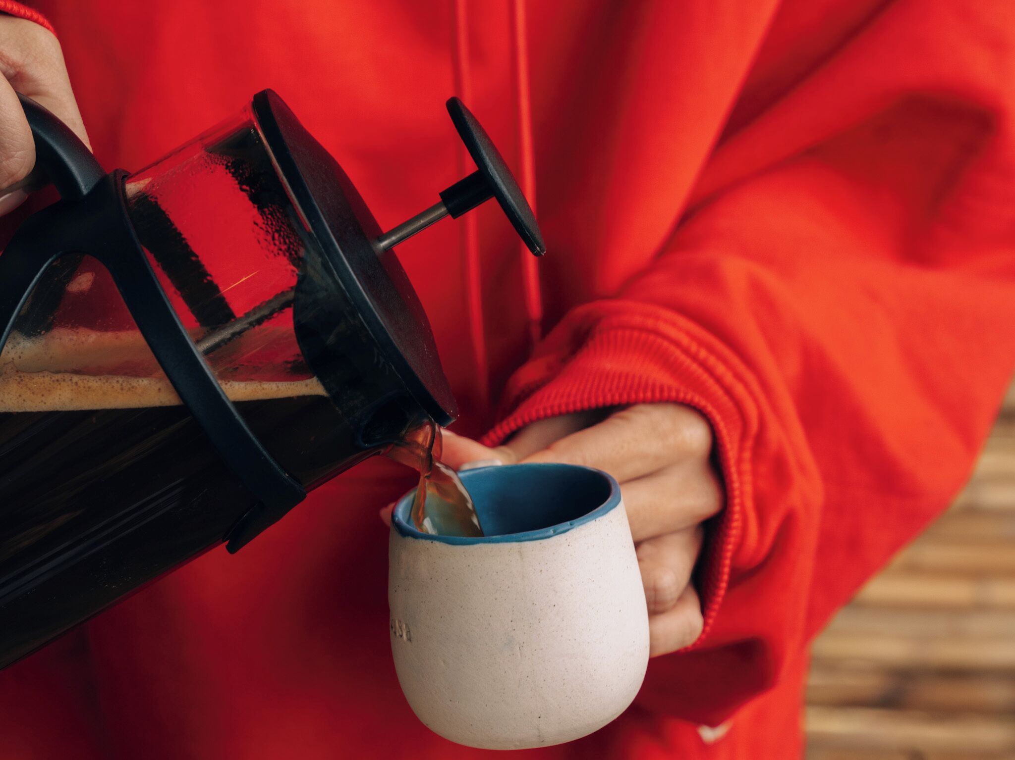 A person in a red hoodie pouring coffee from a black coffee press into a white mug with a blue rim.