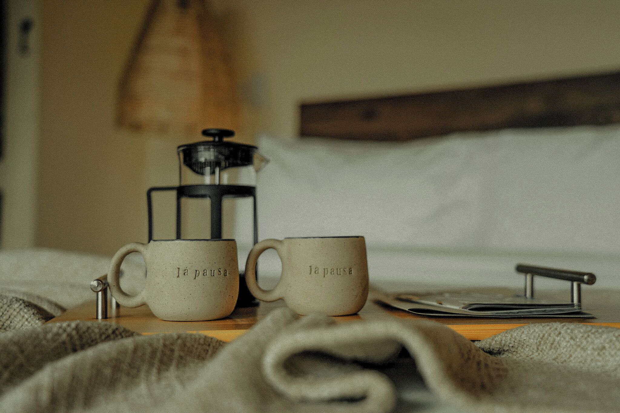 Two coffee cups with text, a coffee press, and a spoon on a table in a room with a bed and a wooden headboard.