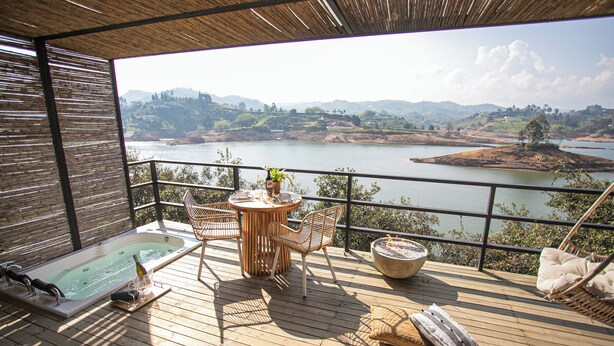 A wooden deck with a hot tub, a table with chairs, and a view of a lake and hills. - La Pausa Hotelbistro (El Peñol)