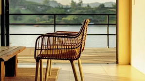A room with a wicker chair, a wooden table, and a view of a lake and hills. - La Pausa Hotelbistro (El Peñol)