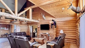 A log cabin interior with a fireplace, leather sofas, wooden tables, and mounted animal heads.