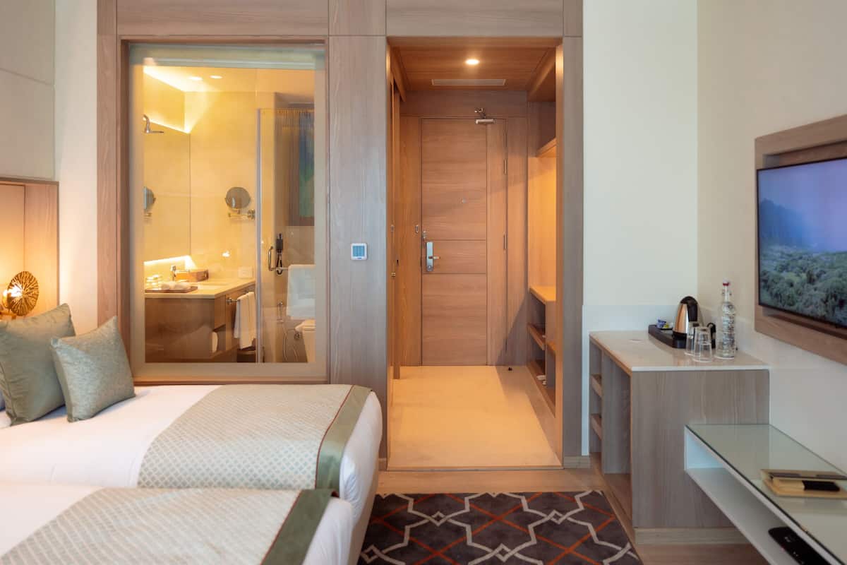 Habitación de hotel con cama, escritorio, televisor y baño visible a través de una puerta de vidrio.