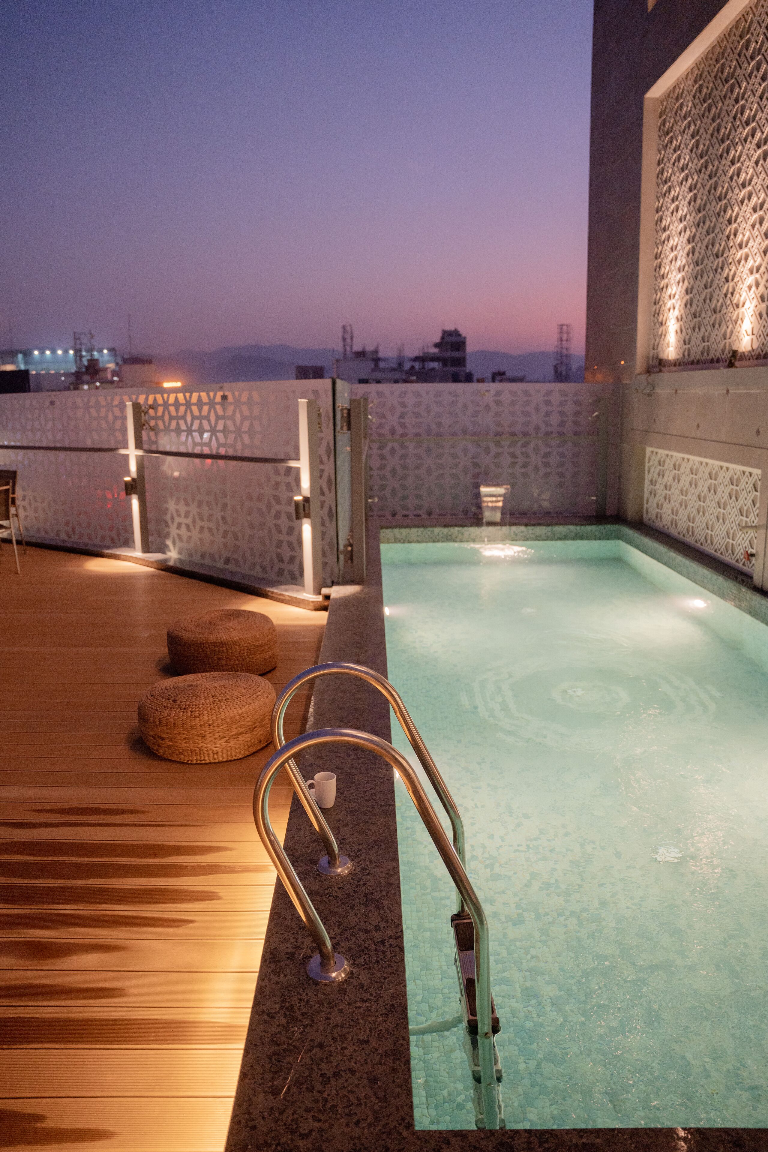 A rooftop pool with a wooden deck, a small seating area, and a view of the cityscape at dusk.