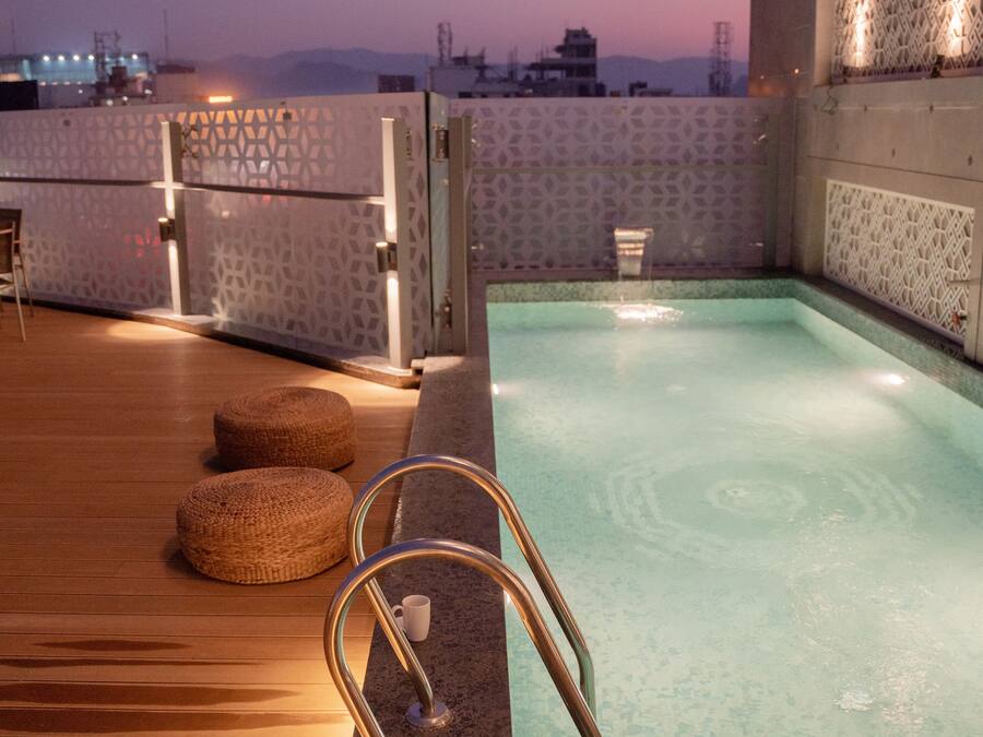 A rooftop pool with a wooden deck, a small seating area, and a view of the cityscape at dusk.