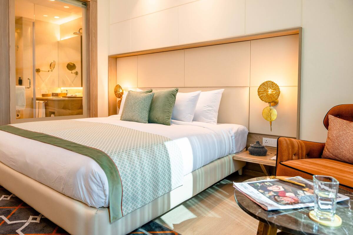 Habitación de hotel con una cama grande, una mesita de noche, una silla y una mesa de vidrio con una revista y un vaso.