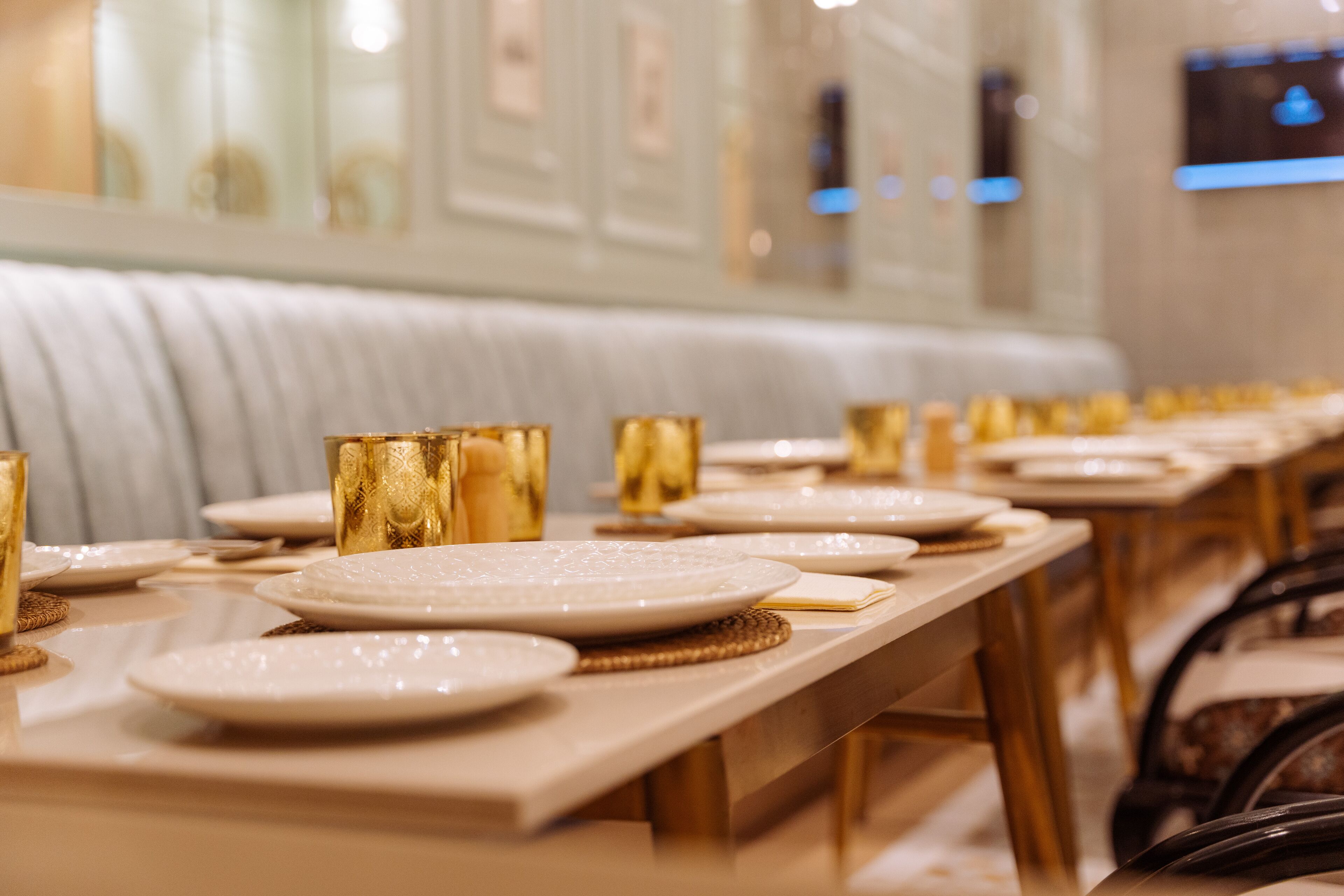 Una larga mesa de comedor dispuesta con platos, cubiertos y vasos dorados, con un banco acolchado al fondo.