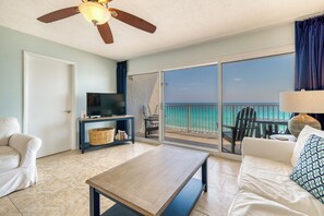 A room with a ceiling fan, a television on a stand, a sofa, a coffee table, and a view of the ocean through a sliding glass door. - C602 | Beachfront with Beach Service, Pool, & WiFi (Miramar Beach)