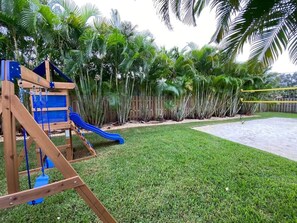 Une structure de jeux en bois avec un toboggan, entourée d’une pelouse bien entretenue et de palmiers.