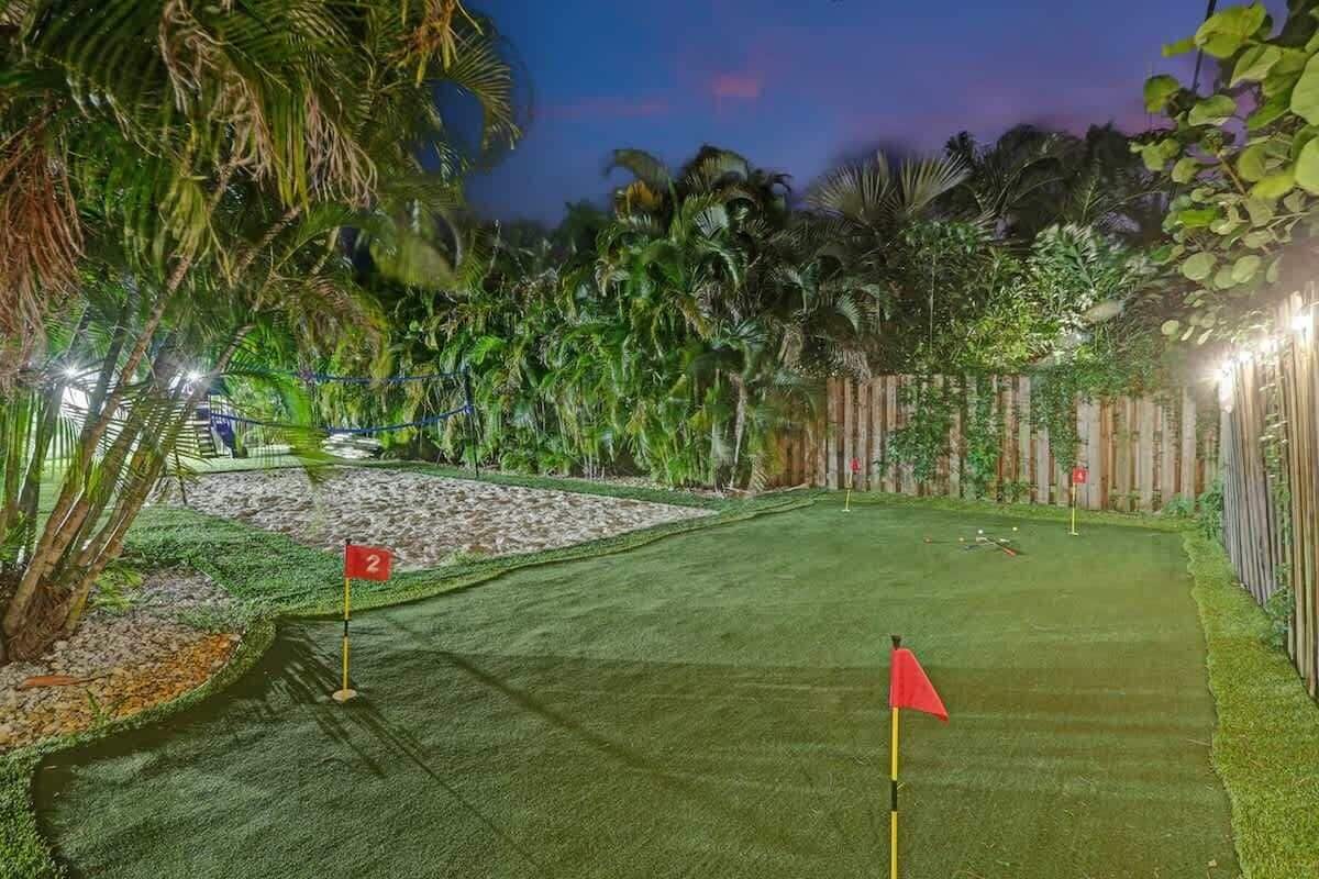 Un campo de golf de césped artificial bien cuidado, con una bandera roja que marca el hoyo, rodeado de exuberante vegetación tropical y una valla de madera.