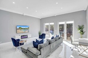 Un salon moderne avec un canapé gris, des fauteuils bleus, un téléviseur à écran plat et une porte en verre donnant sur un espace extérieur.