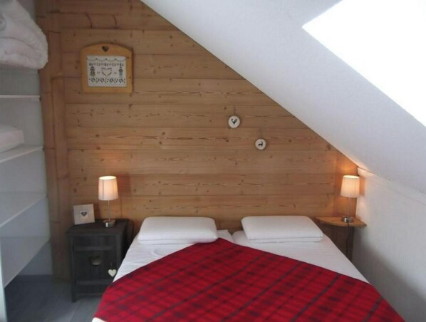 A bedroom with a sloped ceiling, wooden walls, two bedside tables with lamps, a bed with white linens and a red plaid blanket, and a framed picture on the wall.