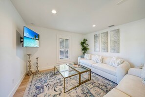 A living room with a white sofa, a glass coffee table, a patterned rug, a TV on the wall, and a window with blinds.