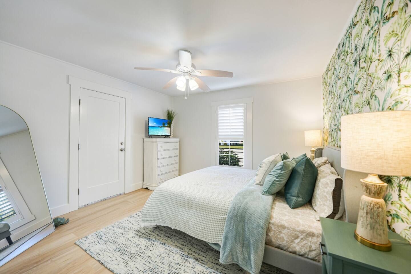 Une chambre avec un lit, un ventilateur de plafond, un téléviseur posé sur une commode, une fenêtre avec des stores et un mur recouvert d’un papier peint aux motifs tropicaux.