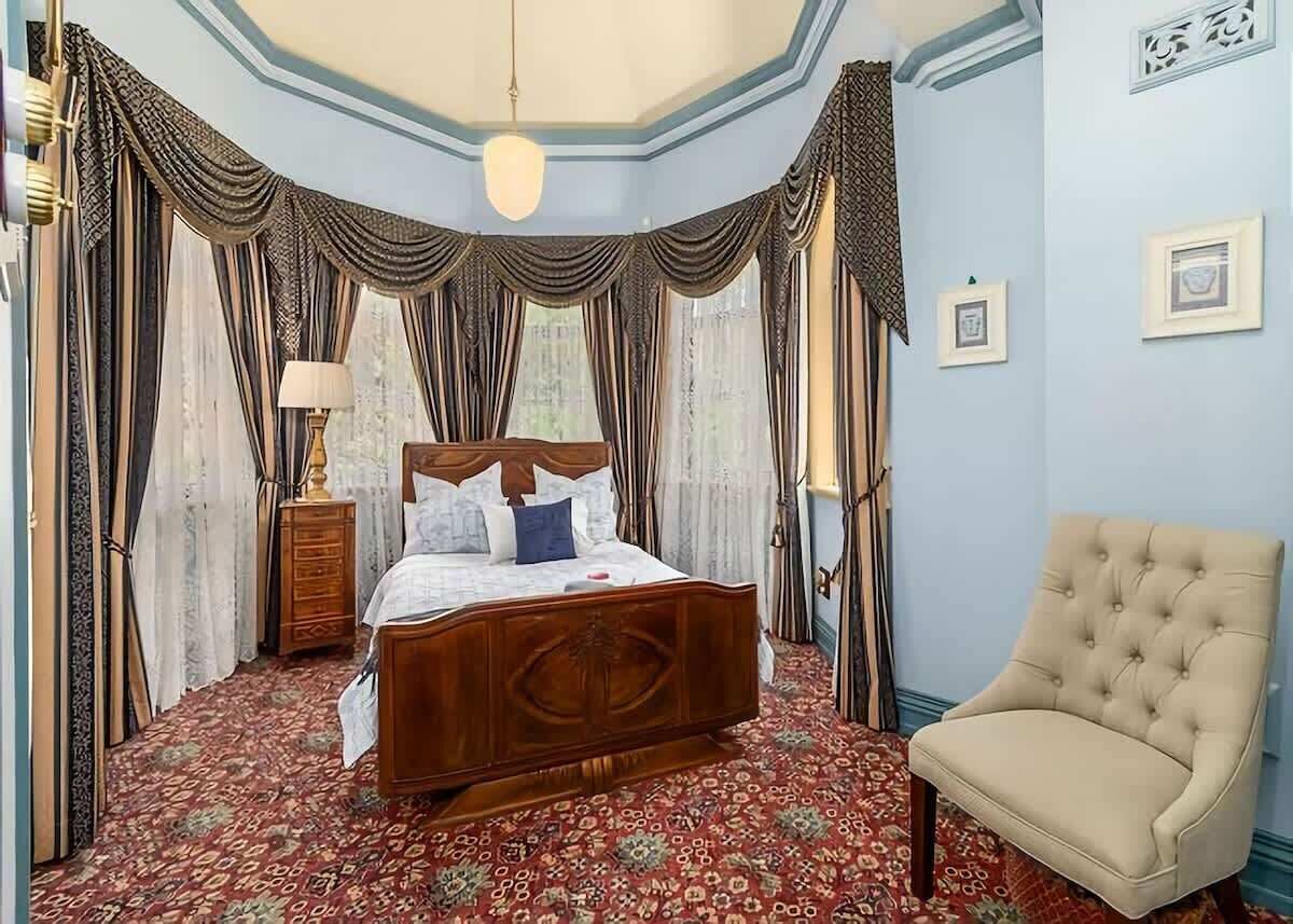Un dormitorio con cama de madera, una silla, cortinas e imágenes enmarcadas en la pared.