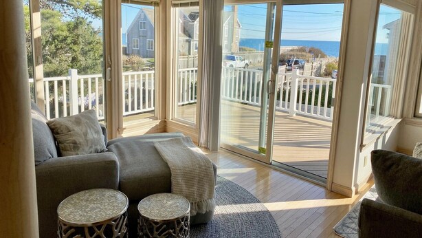 A sunlit room with a large sliding glass door leading to a deck with a white railing, a gray sofa, and two round side tables. - 🌊 BEACHFRONT FAMILY HOME - 2026 Vrbo Rental Of The Year Award! (Yarmouth)