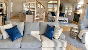 A spacious living area with a grey sofa, a staircase, a fireplace, and a kitchen in the background. - 🌊 BEACHFRONT FAMILY HOME - 2026 Vrbo Rental Of The Year Award! (Yarmouth)